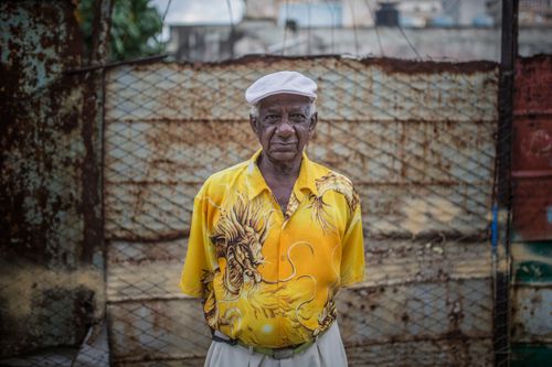 Free Elderly man in a vibrant dragon shirt standing in front of a rustic metal fence in Cuba. Stock Photo