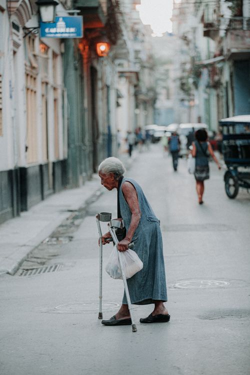 Free An elderly woman with crutches walking down a historic street in Cuba, dressed in a blue dress. Stock Photo