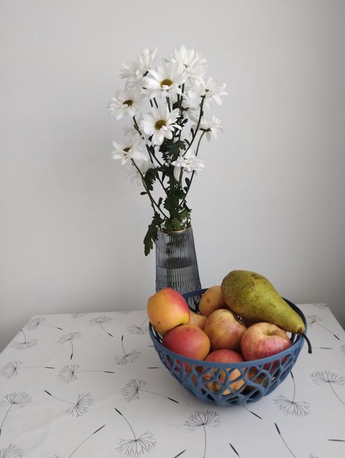 Free Aesthetic arrangement of daisies in vase and colorful fruits in a bowl on a patterned tablecloth. Stock Photo