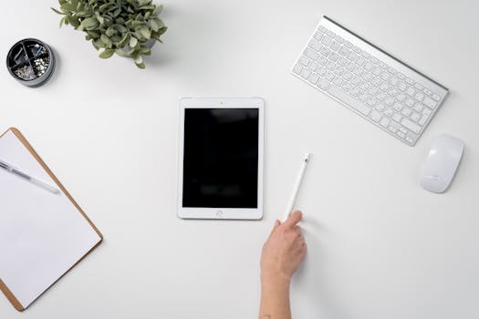 A clean and organized home office setup featuring a tablet, stylus, keyboard, and potted plant.