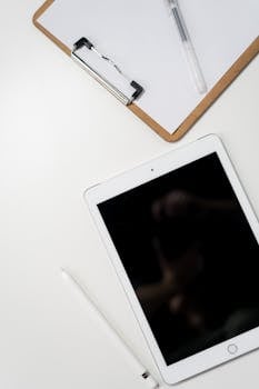 A clean minimalist workspace featuring a tablet, stylus, and clipboard on a white desk.