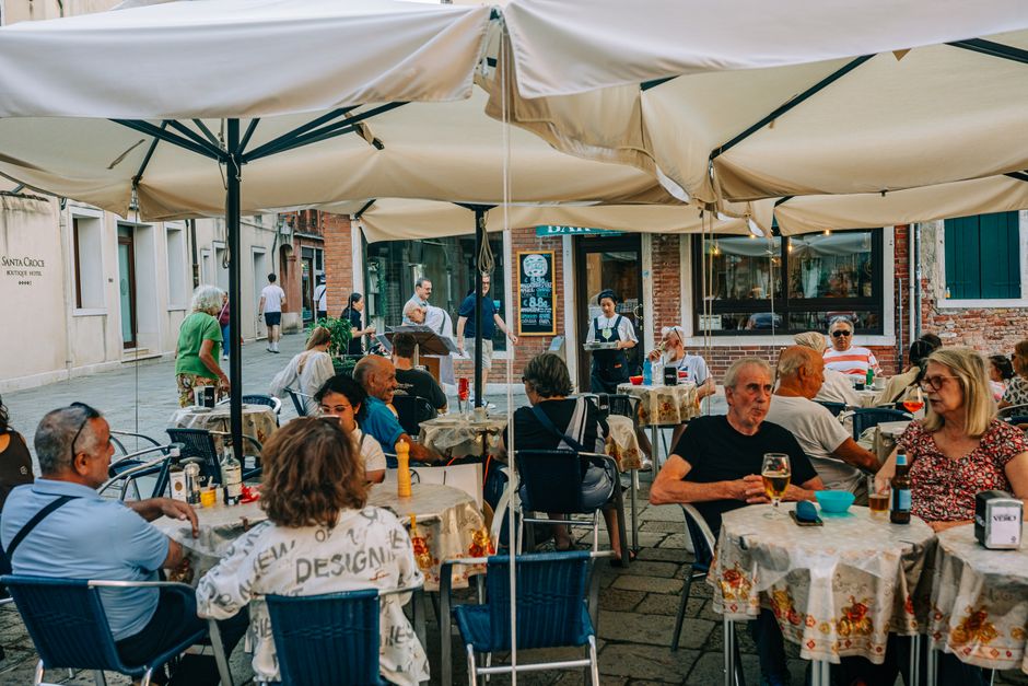 Lively outdoor cafe with people enjoying meals and drinks under umbrellas on a bustling European street.