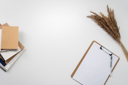 A minimalist home office setup with notebooks, clipboard, and dried flowers on a white background.