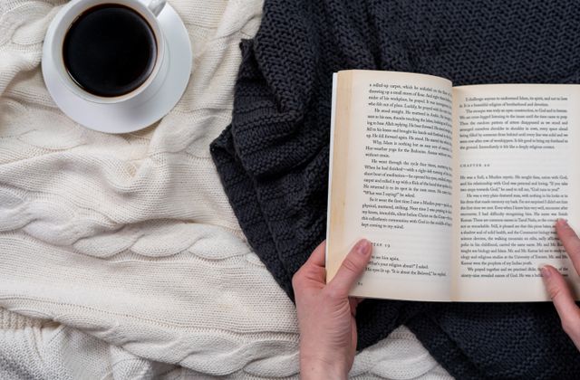 An open book and a cup of coffee resting on a knitted blanket