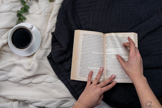 双手捧着一本打开的书，书上放着一杯咖啡，腿上盖着舒适的毯子