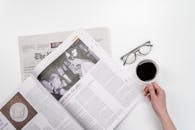 白色桌子上方，俯视视角下，有人正喝着咖啡、拿着杯子阅读报纸