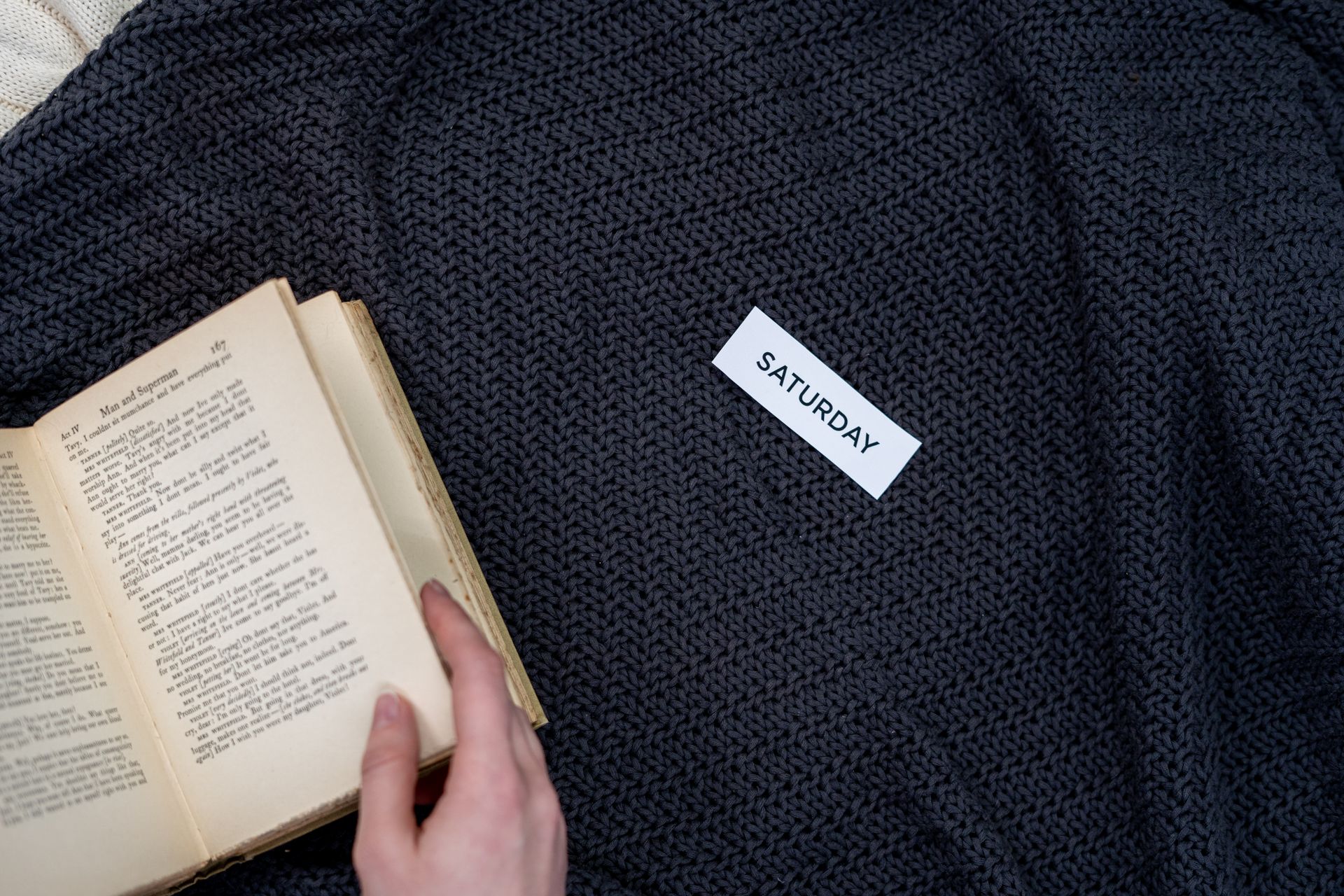 A hand holds an open book on a warm blanket, with Saturday card, evoking leisure.