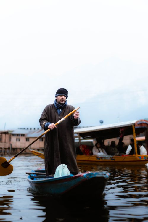 Free A man in traditional clothing paddles a boat on a serene lake, capturing winter's tranquil beauty. Stock Photo