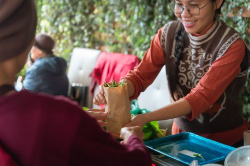 Free Smiling vendor hands paper bag of fresh produce to customer at indoor market stall. Stock Photo