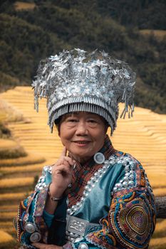 Elderly Miao woman in vibrant traditional attire with rice terraces in Guangxi, China.