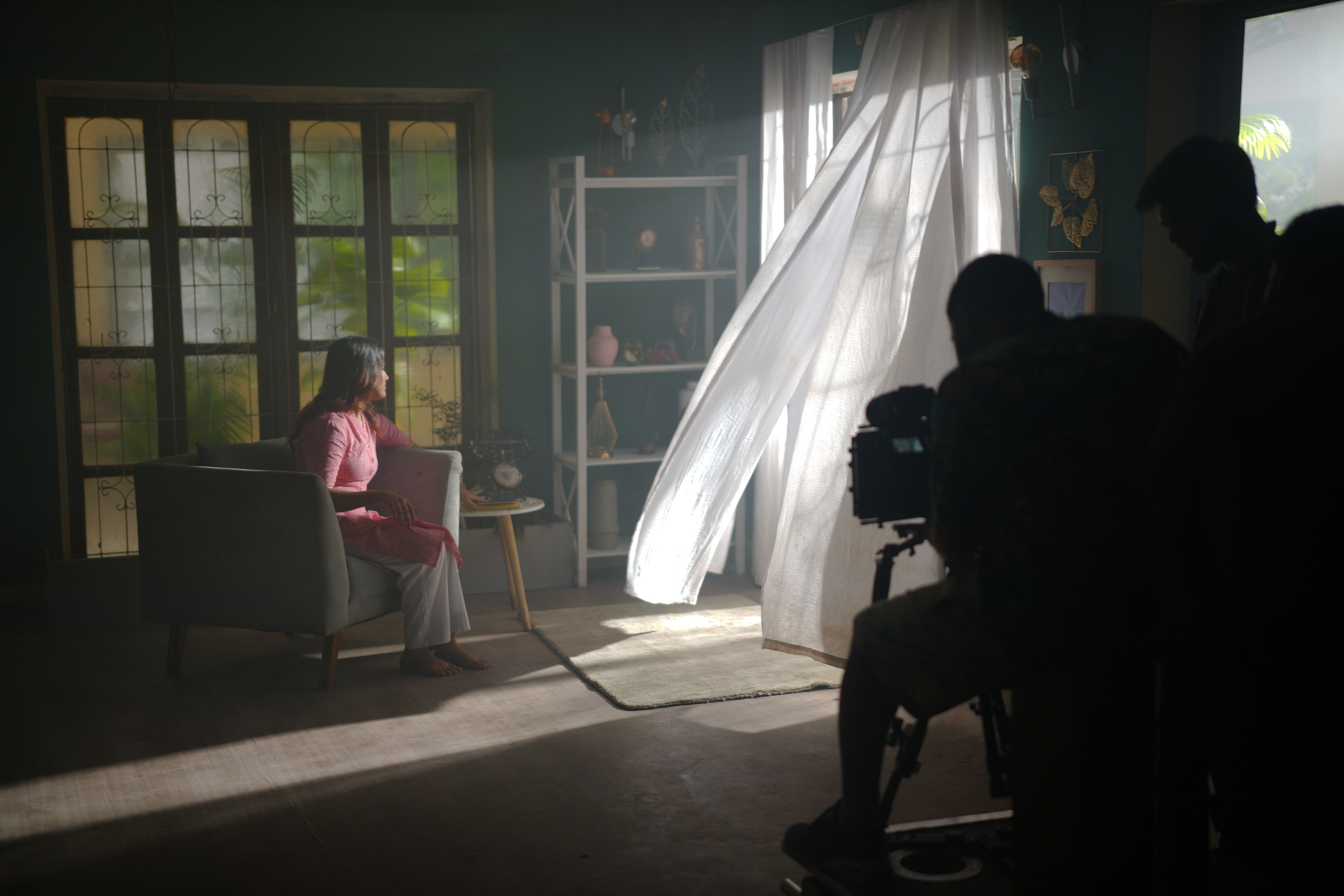 A woman sits in a dimly lit room while curtains billow, creating a cinematic atmosphere.