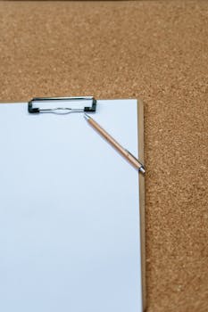 A flatlay of a clipboard with a notepad and pen on a corkboard, ideal for work or planning concepts.