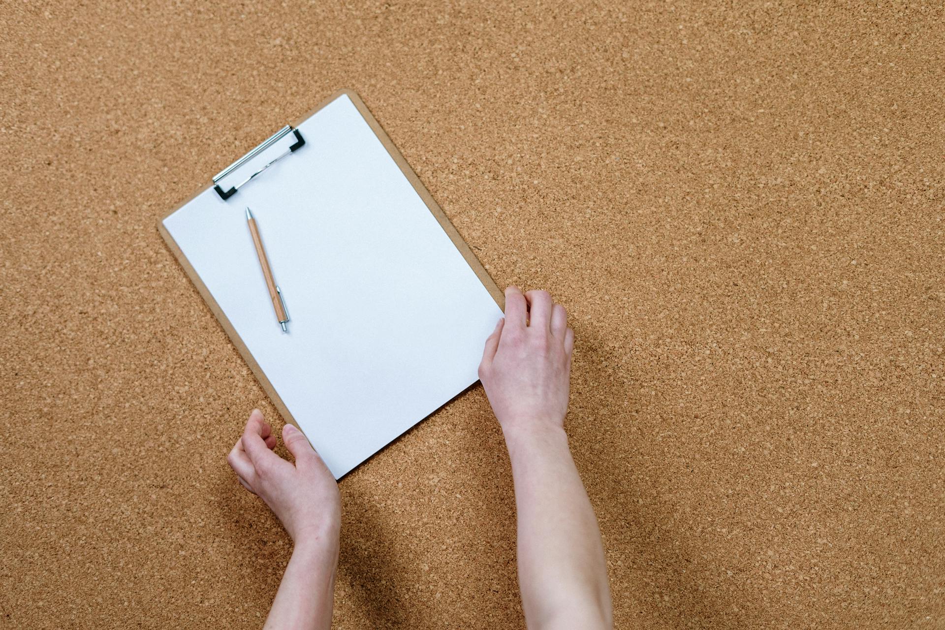 Hands holding a clipboard with paper and pencil on a cork board. Ideal for office or school themes.