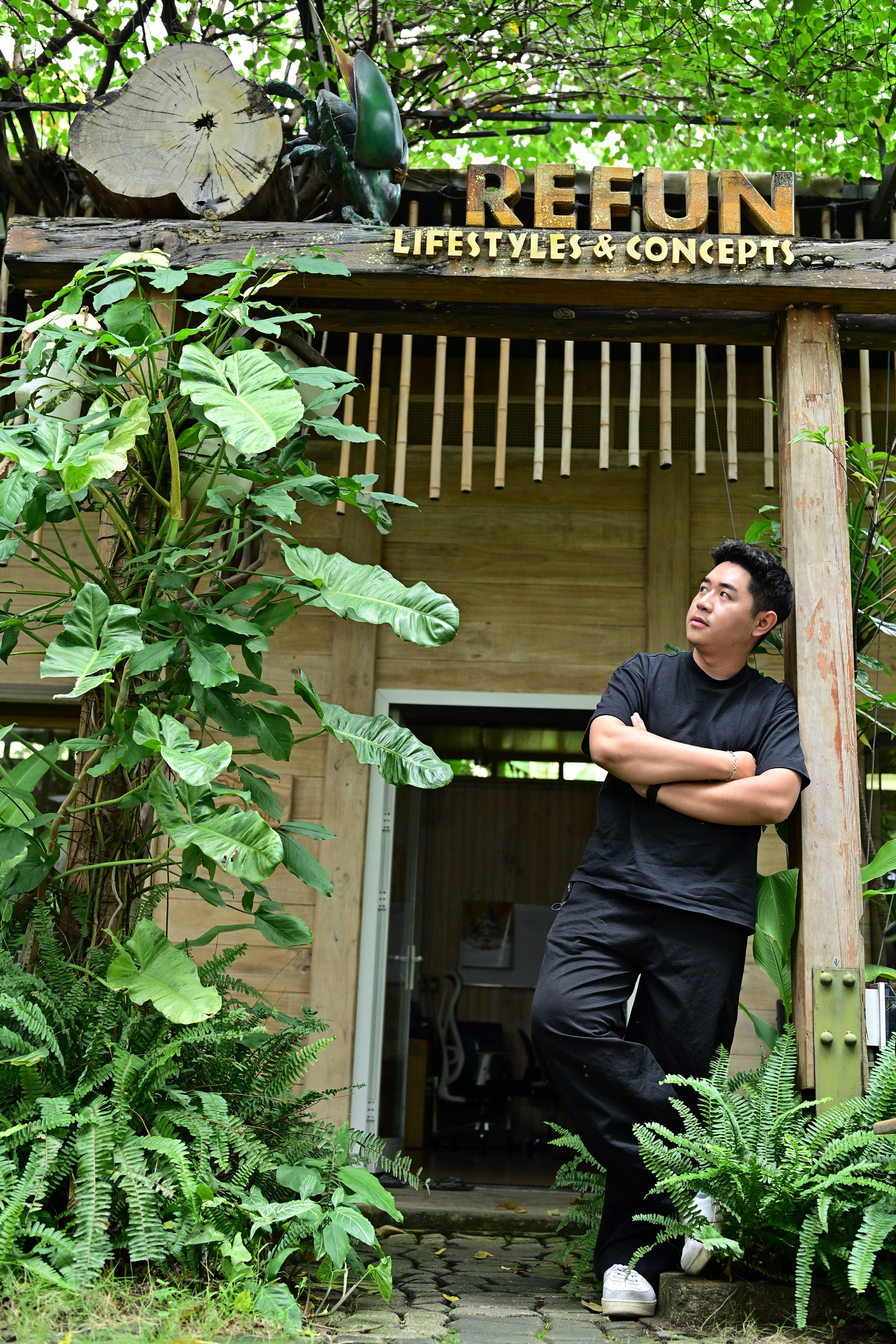 A man leans casually at the entrance of an eco-friendly cafe surrounded by lush greenery.