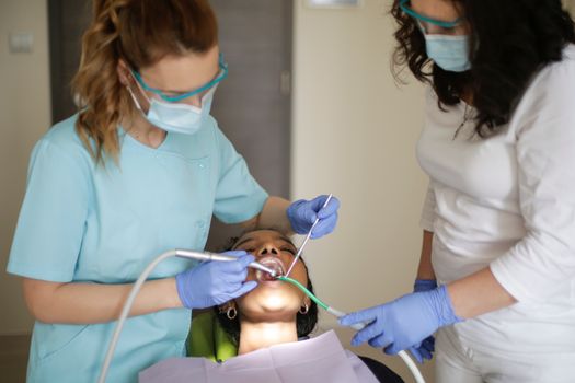 Dentist and assistant in mask uniform and latex gloves treating teeth of black female while working with medical equipment in clinic