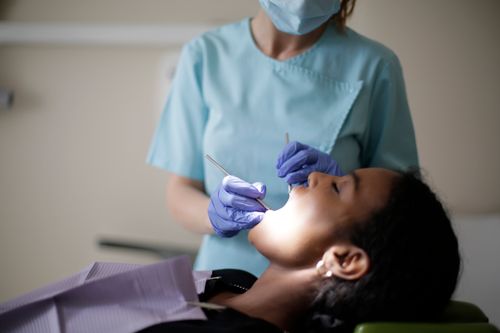 Free Focused female dentist in mask uniform and latex gloves using instruments for examining teeth of African American lady while patient lying with opened mouth and closed eyes Stock Photo