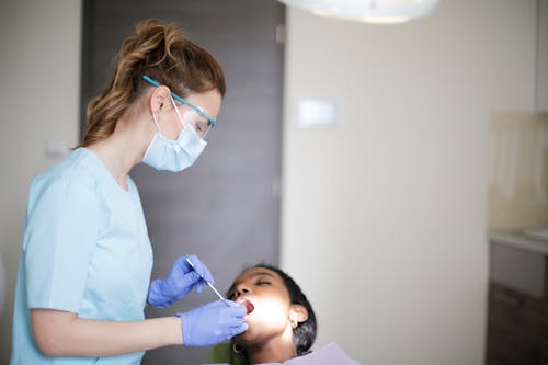 Young female dentist in uniform mask gloves and eyeglasses with dental equipment treating teeth of black patient with closed eyes and opened mouth