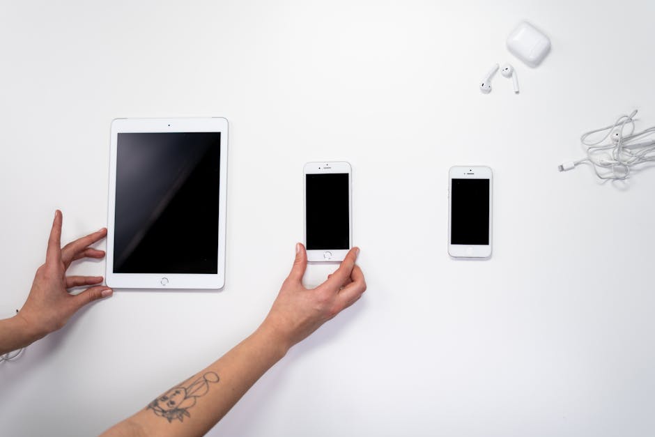 Top view of hands arranging an iPad, iPhones, and earphones on a white surface. Perfect for tech-related imagery.