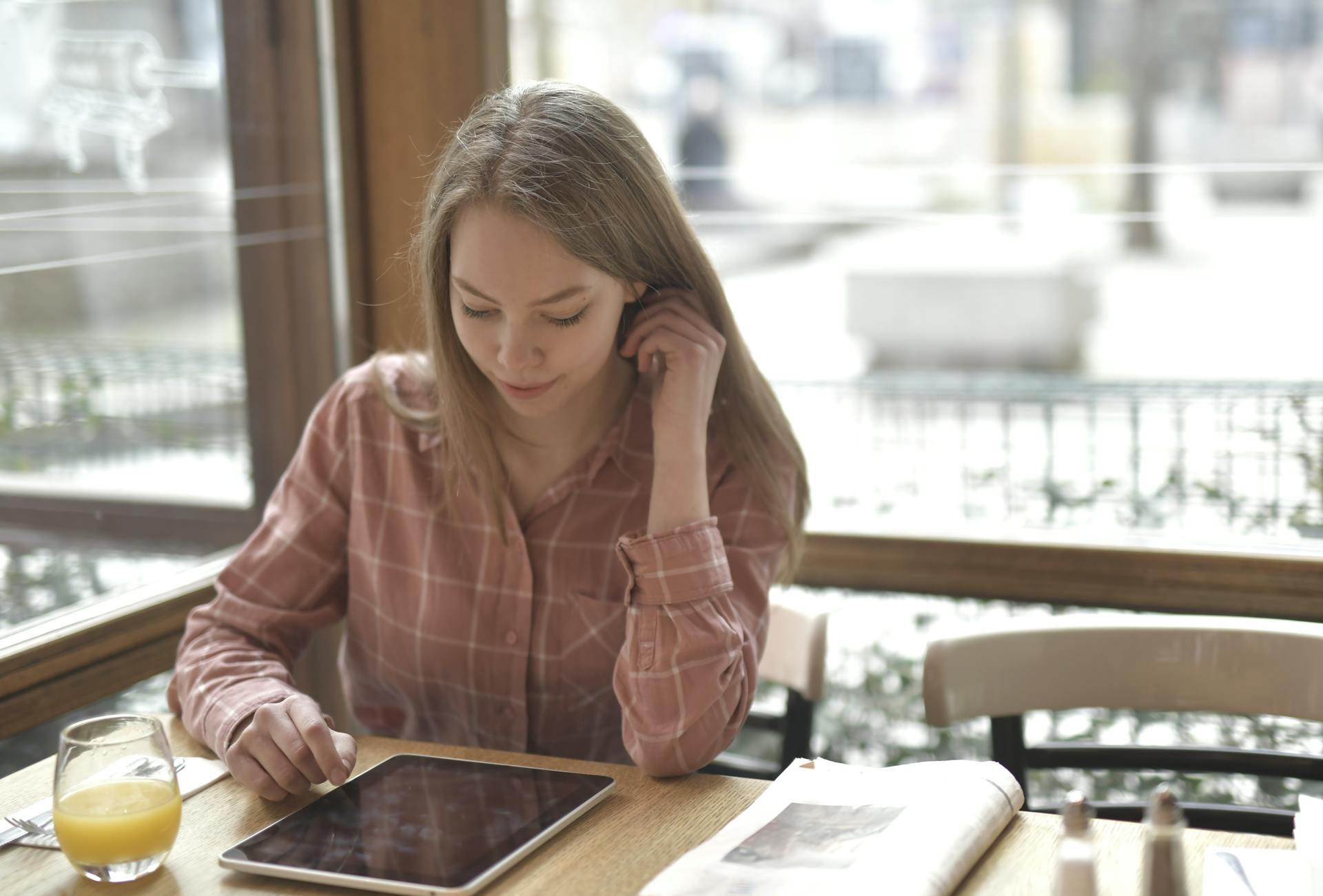 Cheerful concentrated female in checkered shirt sitting at table with glass of fresh juice and newspaper while using tablet