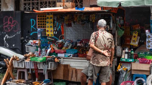 Free Elderly man browsing at a bustling street market stall with various tools and goods displayed. Stock Photo