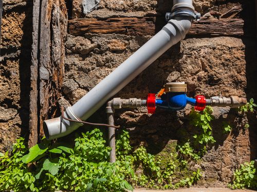 Free A weathered stone wall with a water pipe, meter, and greenery, depicting rustic charm. Stock Photo