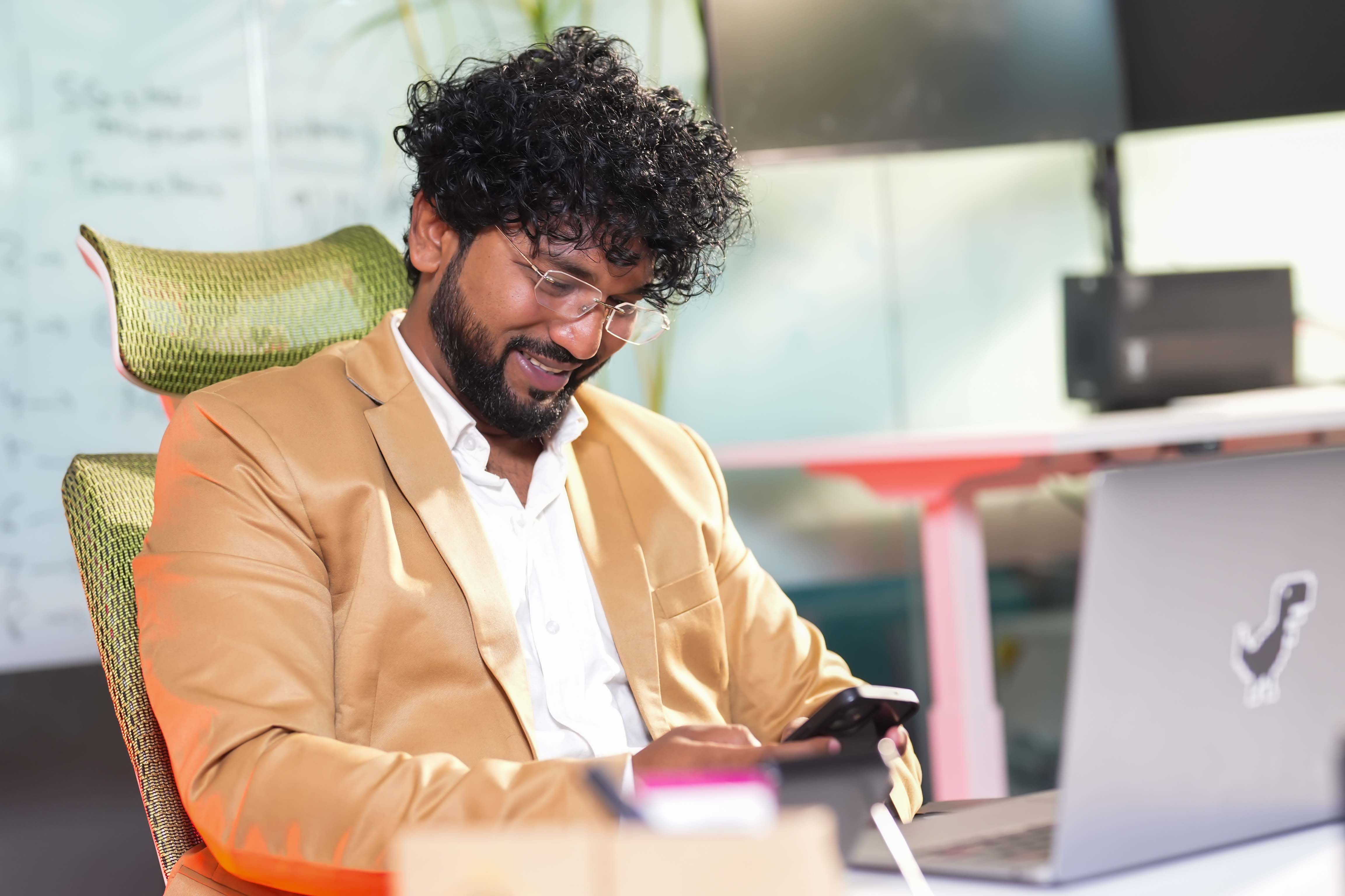 Confident businessman in an office setting smiling at his smartphone while seated at a desk with a laptop.