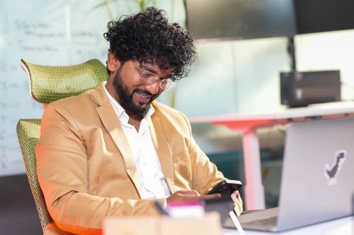 Free Confident businessman in an office setting smiling at his smartphone while seated at a desk with a laptop. Stock Photo