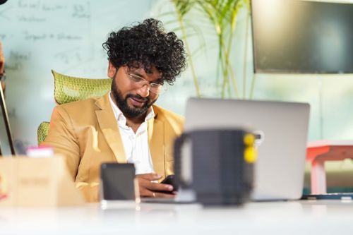 Free A professional man in a beige suit using a smartphone at a modern office desk with a laptop. Stock Photo