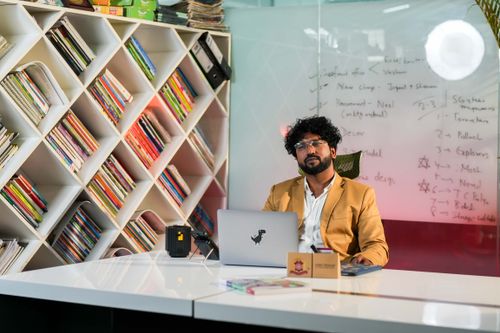 Free Businessman sitting at desk in a modern office with a bookshelf and whiteboard in the background. Stock Photo
