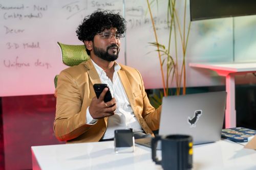 Free Professional man in an office setting using smartphone and laptop, surrounded by workspace essentials. Stock Photo