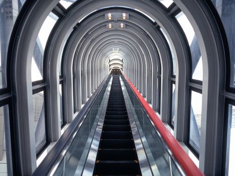 Modern glass tunnel with escalator leading upwards, futuristic architectural design.