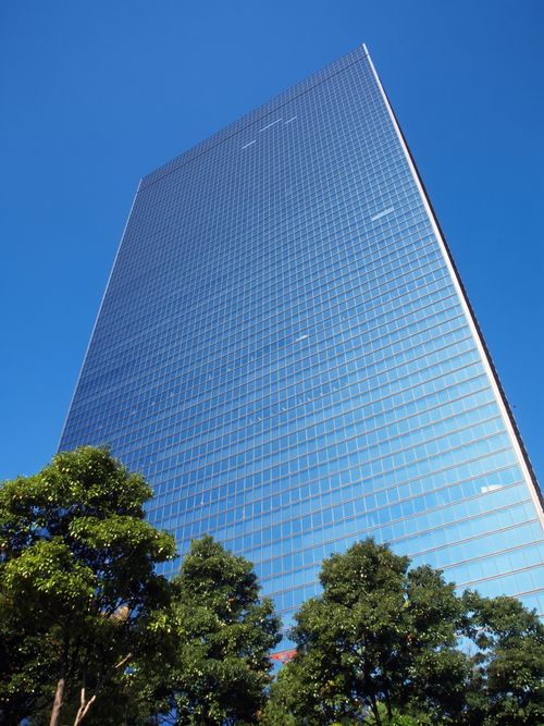 Free Tall glass skyscraper with trees beneath, set against a clear blue sky. Stock Photo
