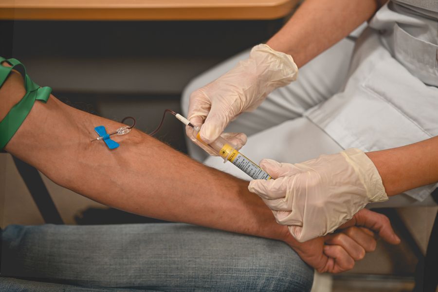 Gloved hands fill a blood tube from a patient's arm, a green tourniquet above the elbow
