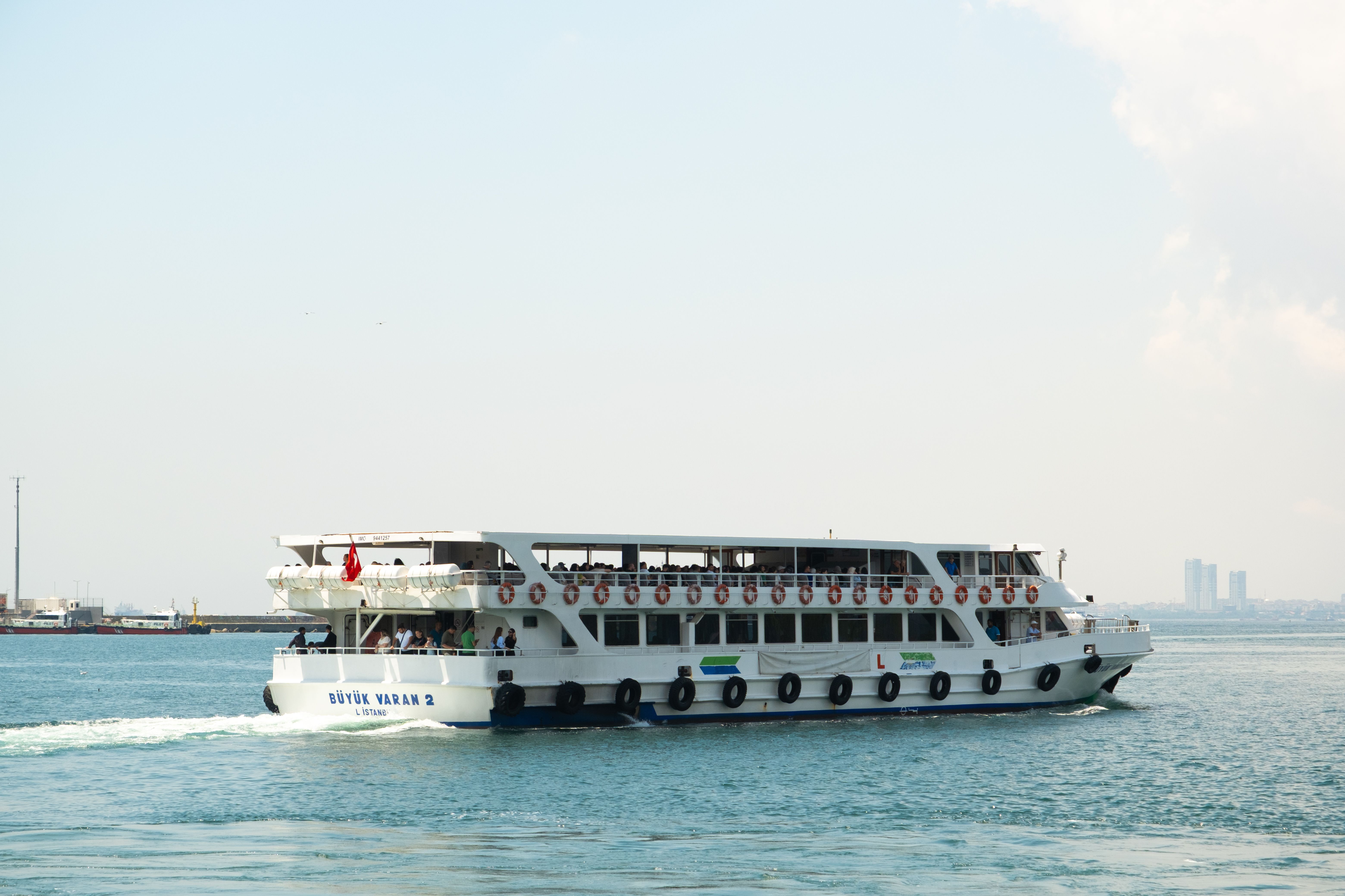 A passenger ferry cruises on a calm sea with a city skyline in the background under a clear sky.