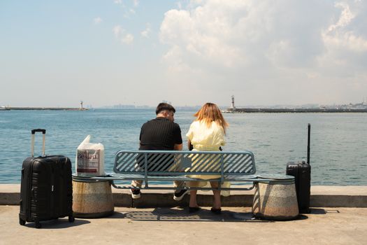 A couple seated on a seafront bench with luggage, enjoying a sunny day by the ocean.