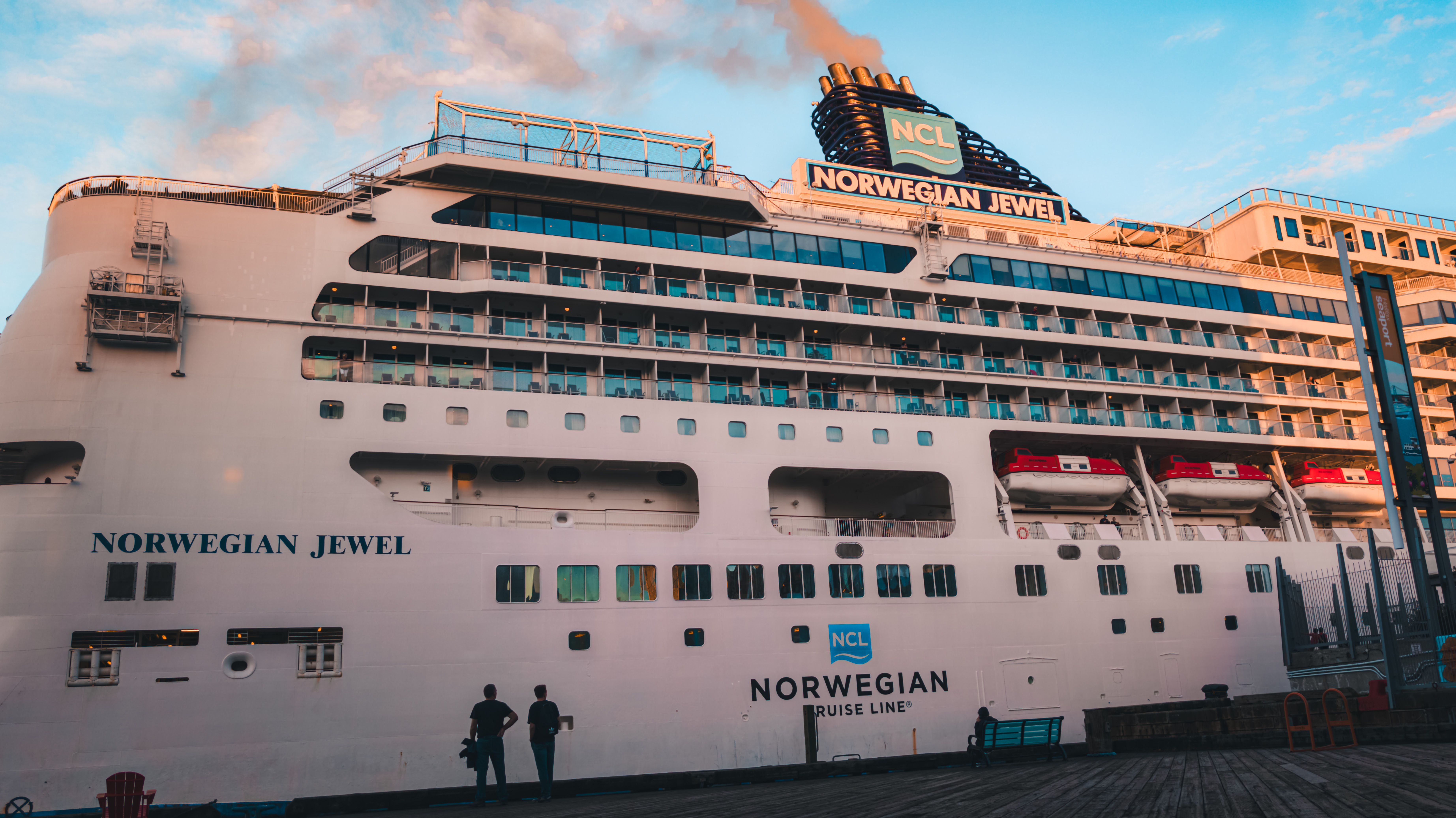Side view of Norwegian Jewel cruise ship docked, basking in sunset light with passengers on shore.