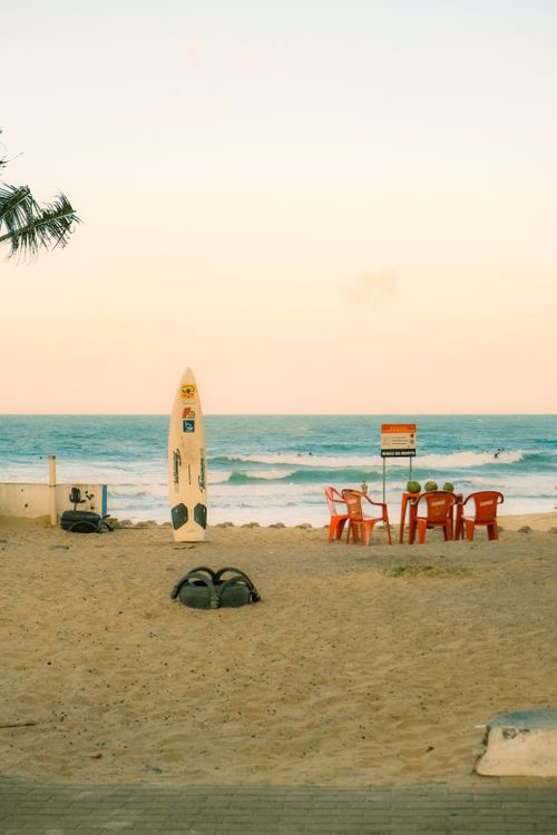 Free A serene beach scene in RN, Brazil with a surfboard, red chairs, and ocean view at sunset. Stock Photo