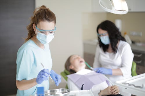 Dentist on mask eyeglasses uniform and gloves choosing material for treating teeth of patient while assistant holding pipe in mouth of patient
