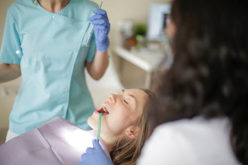 Free Crop faceless female dentist in uniform holding mouth mirror while preparing for inspecting female patient teeth with assistant holding tube suction in patient mouth Stock Photo