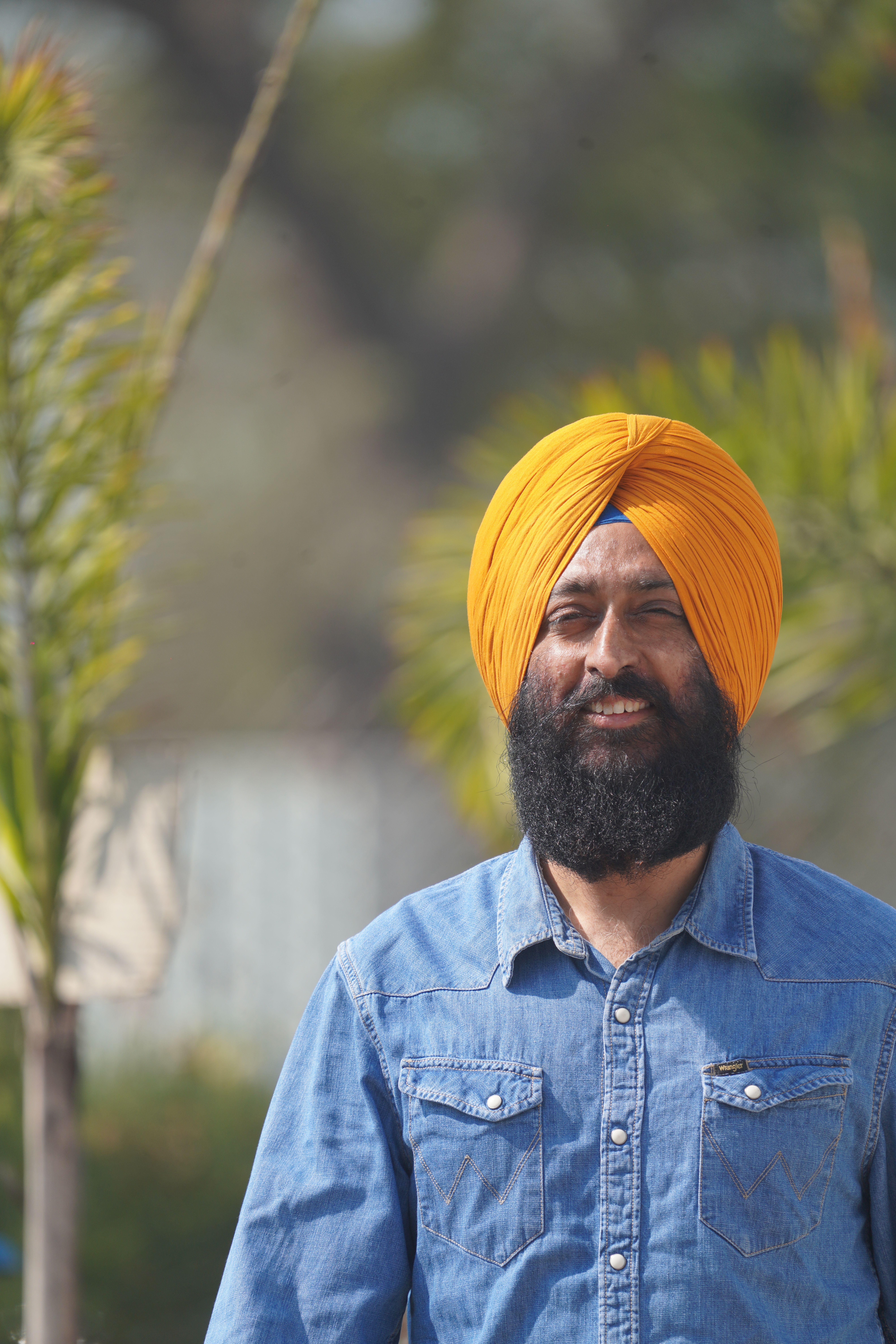 A smiling man wearing an orange turban and denim shirt poses outdoors in a sunny setting.