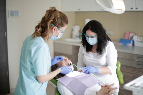 Side view of female dentist in mask wearing blue uniform with latex gloves and protective glasses and assistant using dental tools for examining teeth of female patient lying in dental chair
