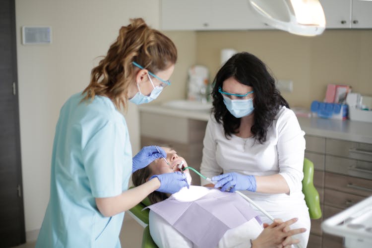 Female Dentist In Mask And Assistant Using Dental Tools For Examining Teeth Of Female Patient