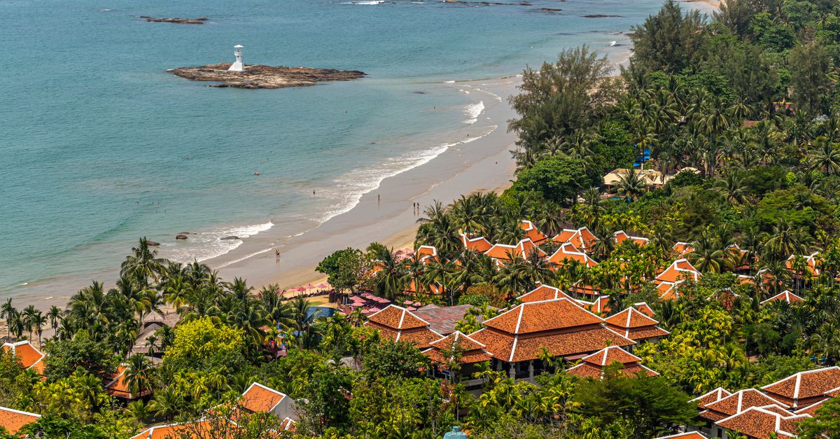 A picturesque beachfront village with terracotta roofs, palm trees, and a distant lighthouse.