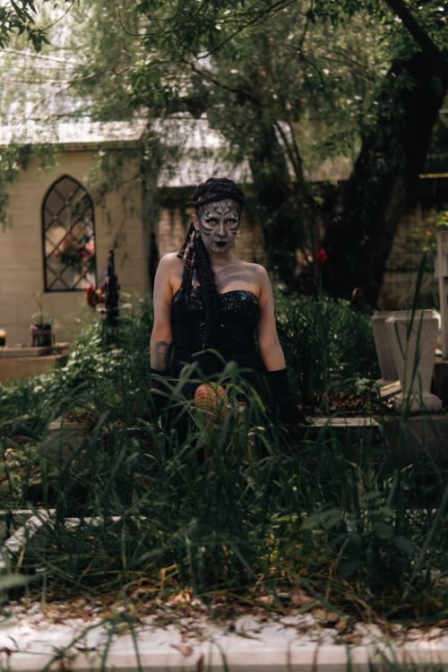 Free A woman in gothic attire with artistic makeup stands in a dimly lit forest setting, evoking mystery and fantasy. Stock Photo