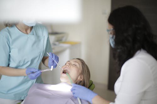 Crop faceless female dentist in blue uniform and latex gloves inserting cotton tampon to patient mouth while working with assistant and preparing patient for dental treatment