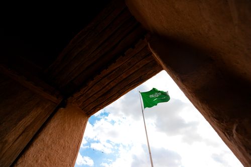 Free Saudi Arabian flag waves against blue sky amid traditional architecture. Stock Photo