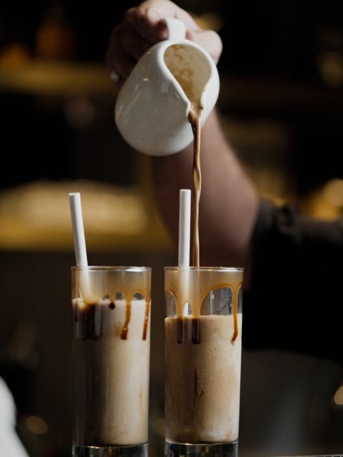 Free Close-up of a barista pouring iced coffee with caramel drizzle in a dimly lit cafe setting. Stock Photo