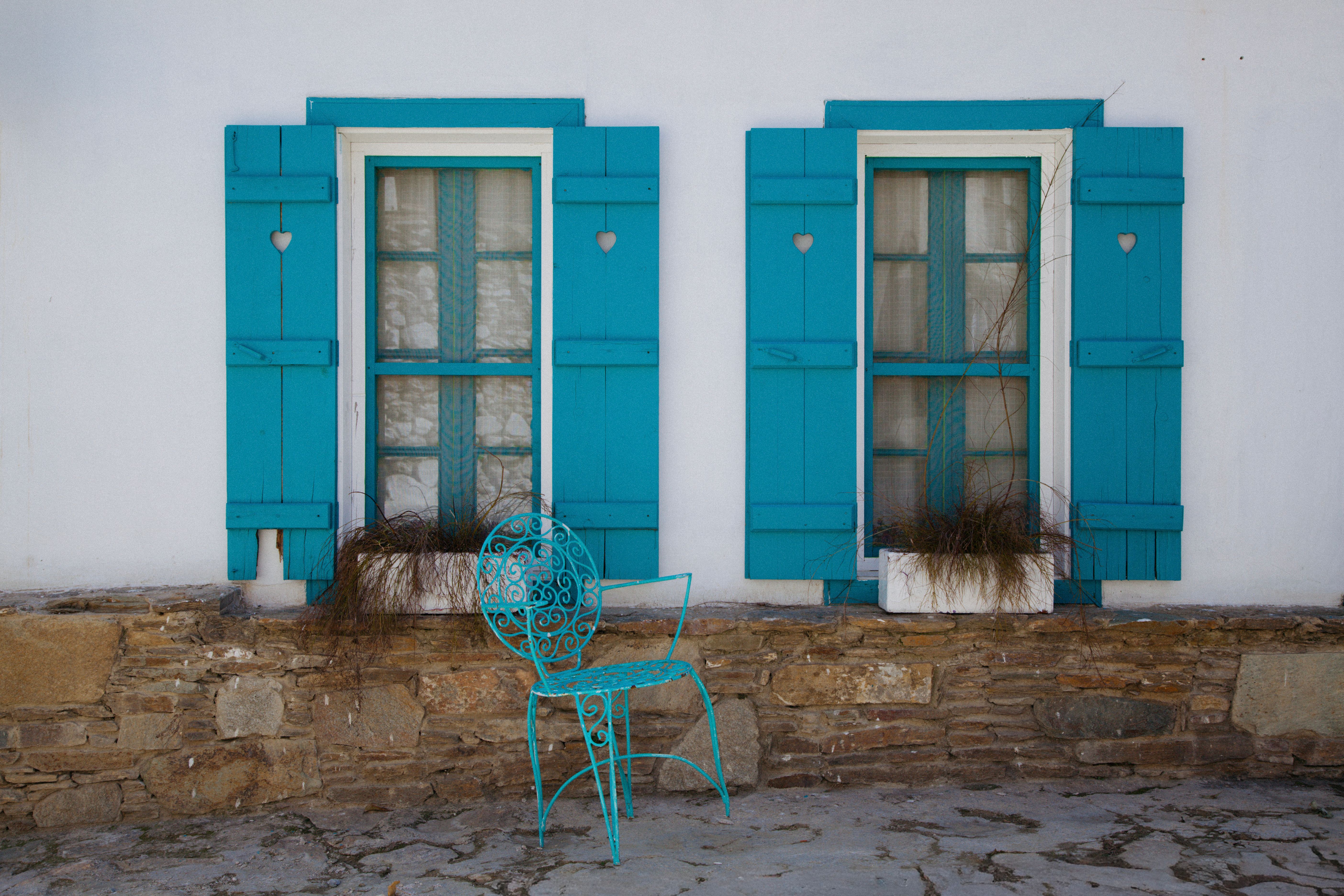 Bright blue shutters and rustic chair against a stone wall exude Mediterranean charm.