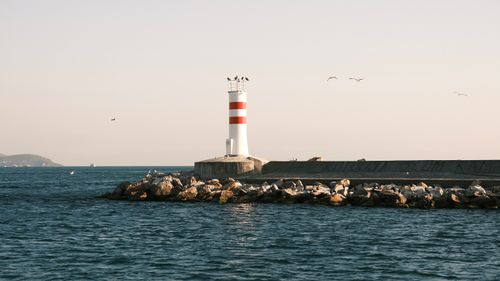 Free Peaceful scene of a lighthouse on rocky shore with tranquil ocean waves at sunset. Stock Photo