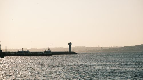 Free A tranquil view of a lighthouse by the sea with boats and gentle waves at sunset. Stock Photo